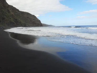 La arena negra mojada de la playa de Nogales reflejando el cielo, con el acantilado y las olas al fondo